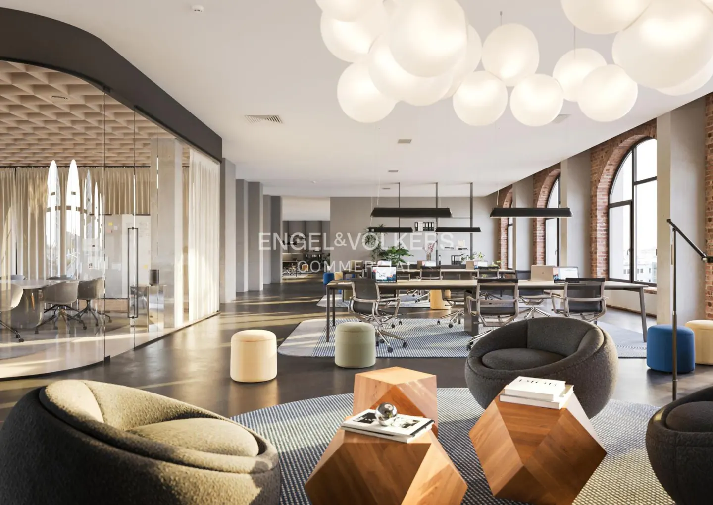 Open office space with lounge area, desks, and large windows. Modern design with gray, beige, and wood tones. Overhead lighting features globe pendants.
