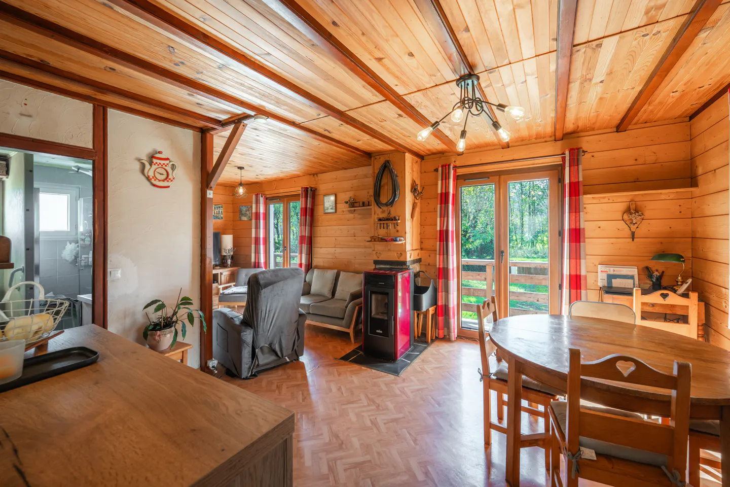 A cozy cabin interior with wood walls, a red stove, and a round wooden table with chairs. A gray sofa and recliner are visible in the background.