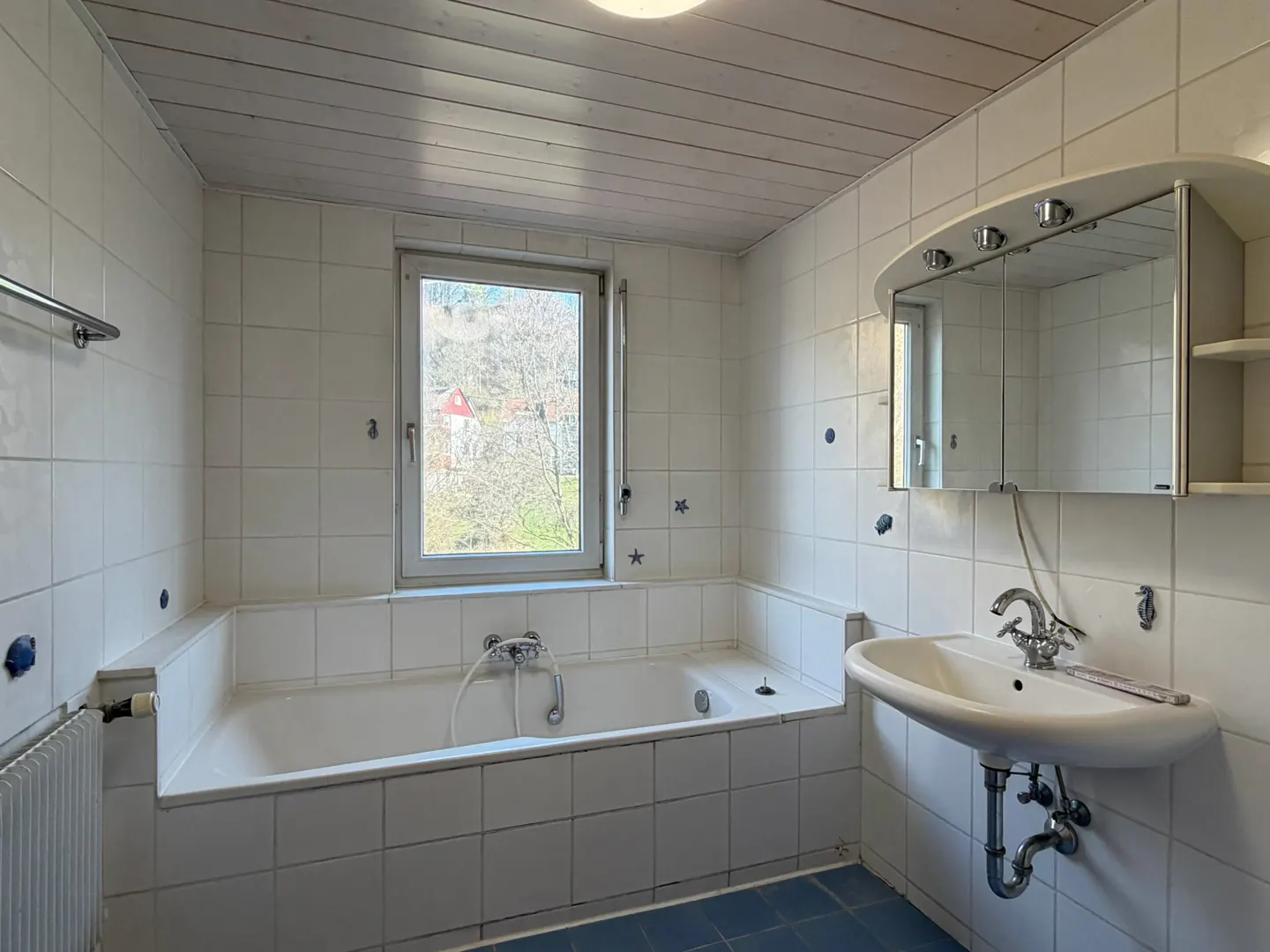 Bright bathroom with white tile walls, blue tile floor, and a window view of trees. A white sink and mirror are on the right.