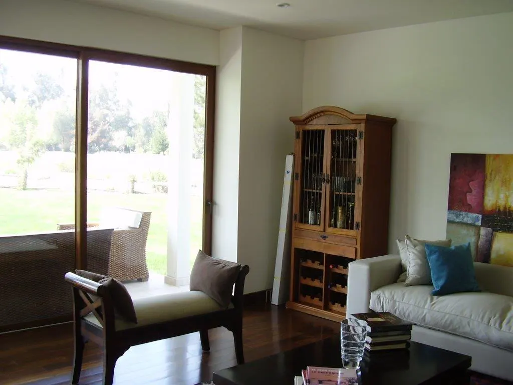 Living room with wood floors, white walls, and large windows overlooking a green lawn. A wooden cabinet, sofa, and bench furnish the space.