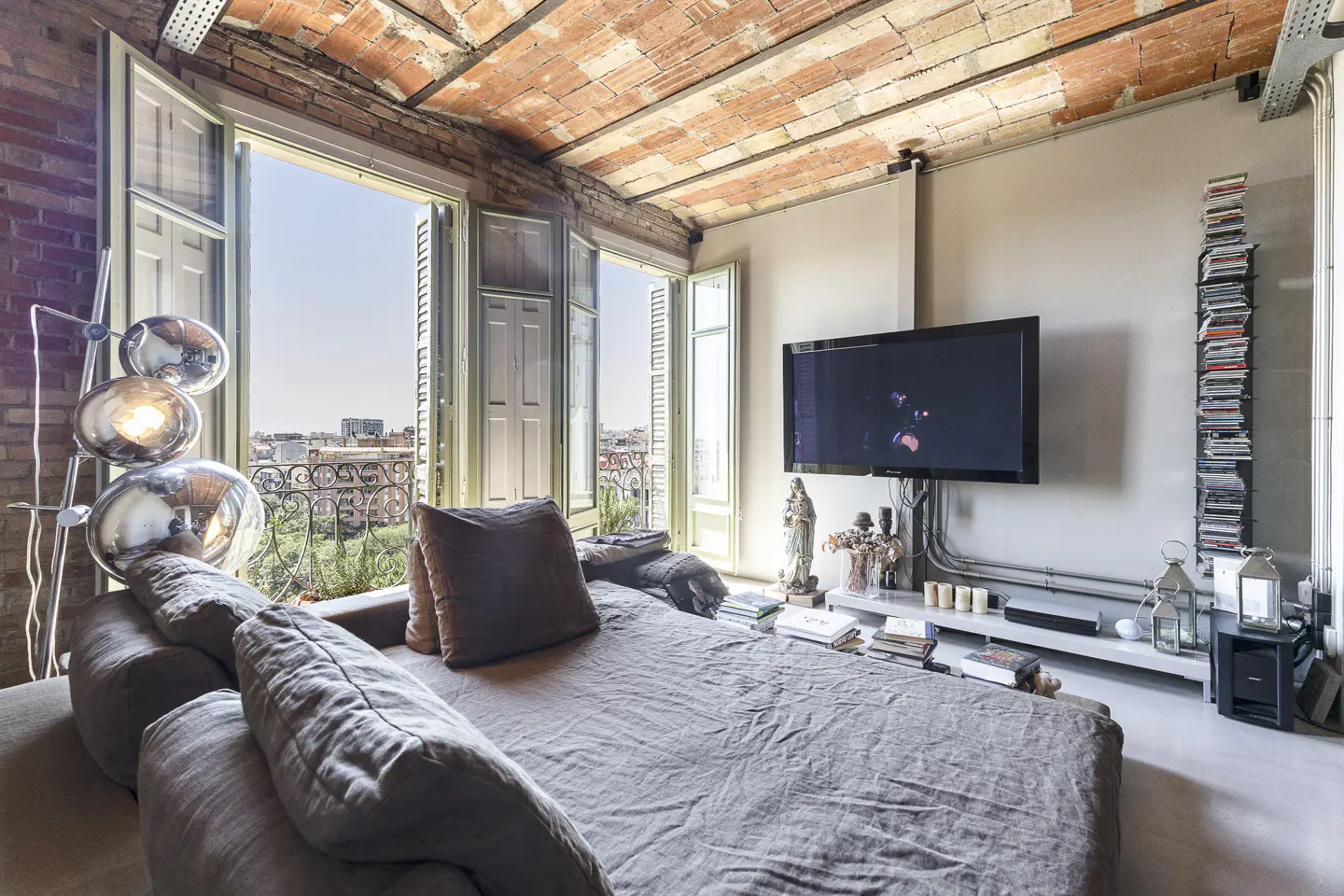 Living room with exposed brick, open windows to a balcony, and a large gray sofa. A TV hangs on the wall with a shelf below.