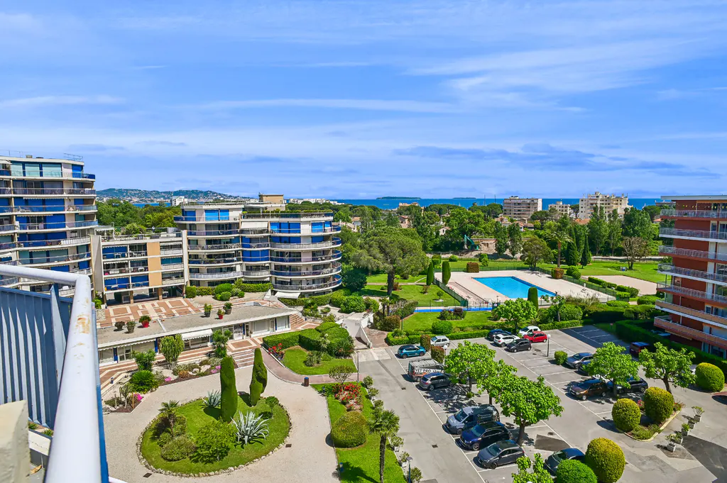 Aerial view of a residential complex with blue-trimmed buildings, a pool, parking lot, and manicured gardens under a blue sky.