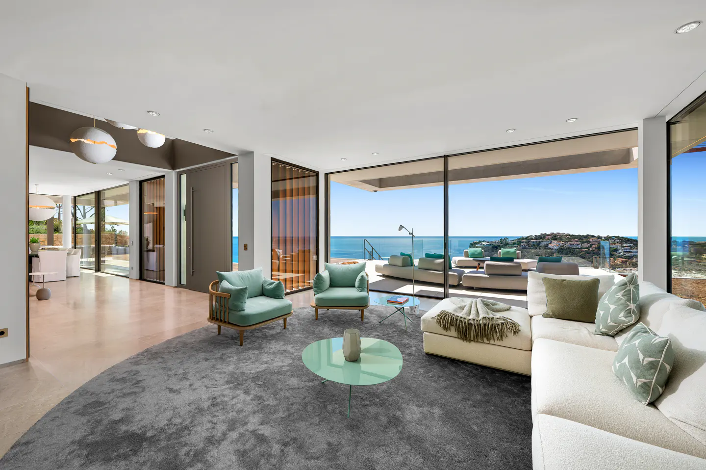 Bright living room with ocean view. Mint chairs, white sofa, and a grey rug. Large windows overlook the ocean.