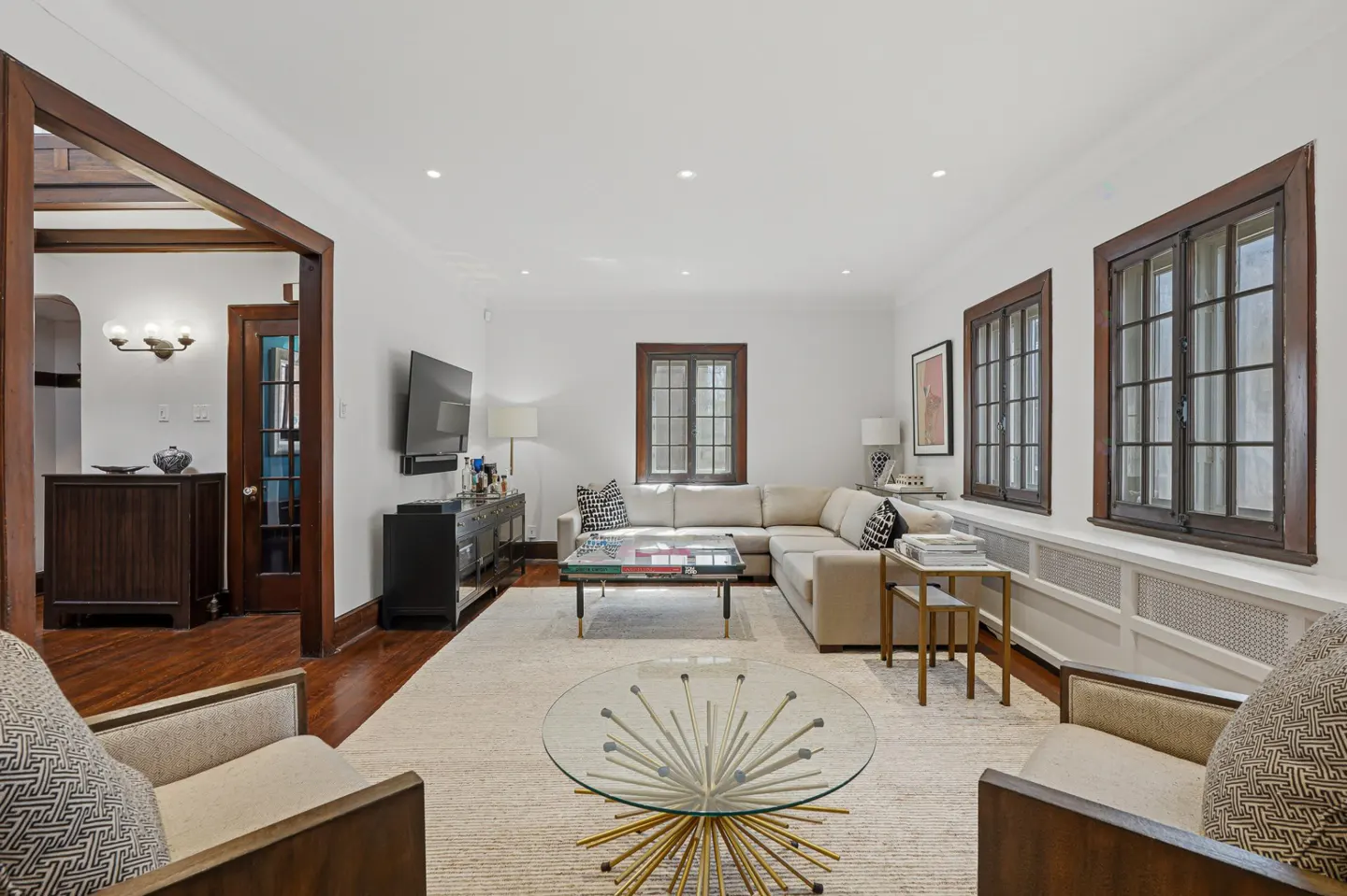 Bright living room with white walls, dark wood trim, and a beige sectional sofa. A glass coffee table sits on a light rug.