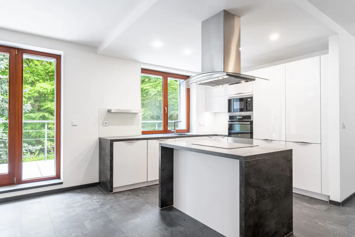 Bright, modern kitchen with white cabinets, dark countertops, and stainless steel hood. Large windows offer a view of lush greenery.