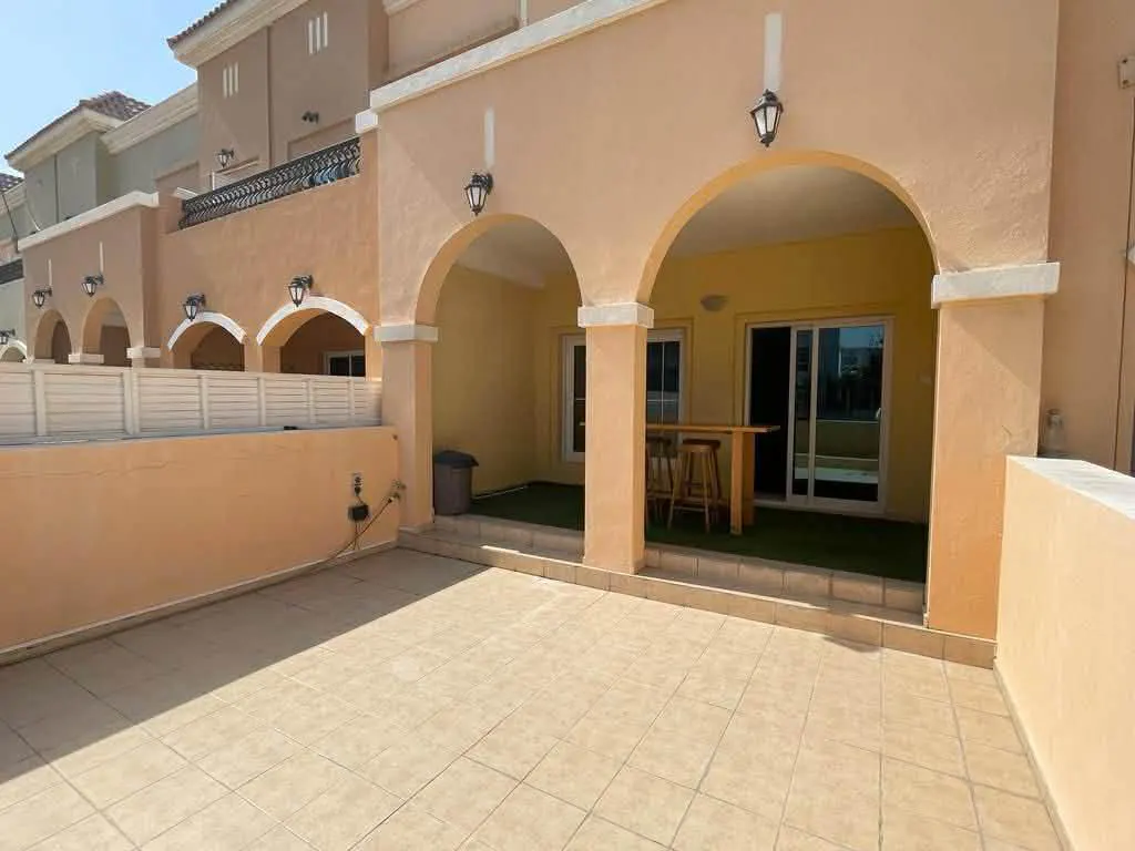 Outdoor patio with beige tile flooring, arched doorways, and a wooden bar table with stools. The building is peach-colored with black sconces.