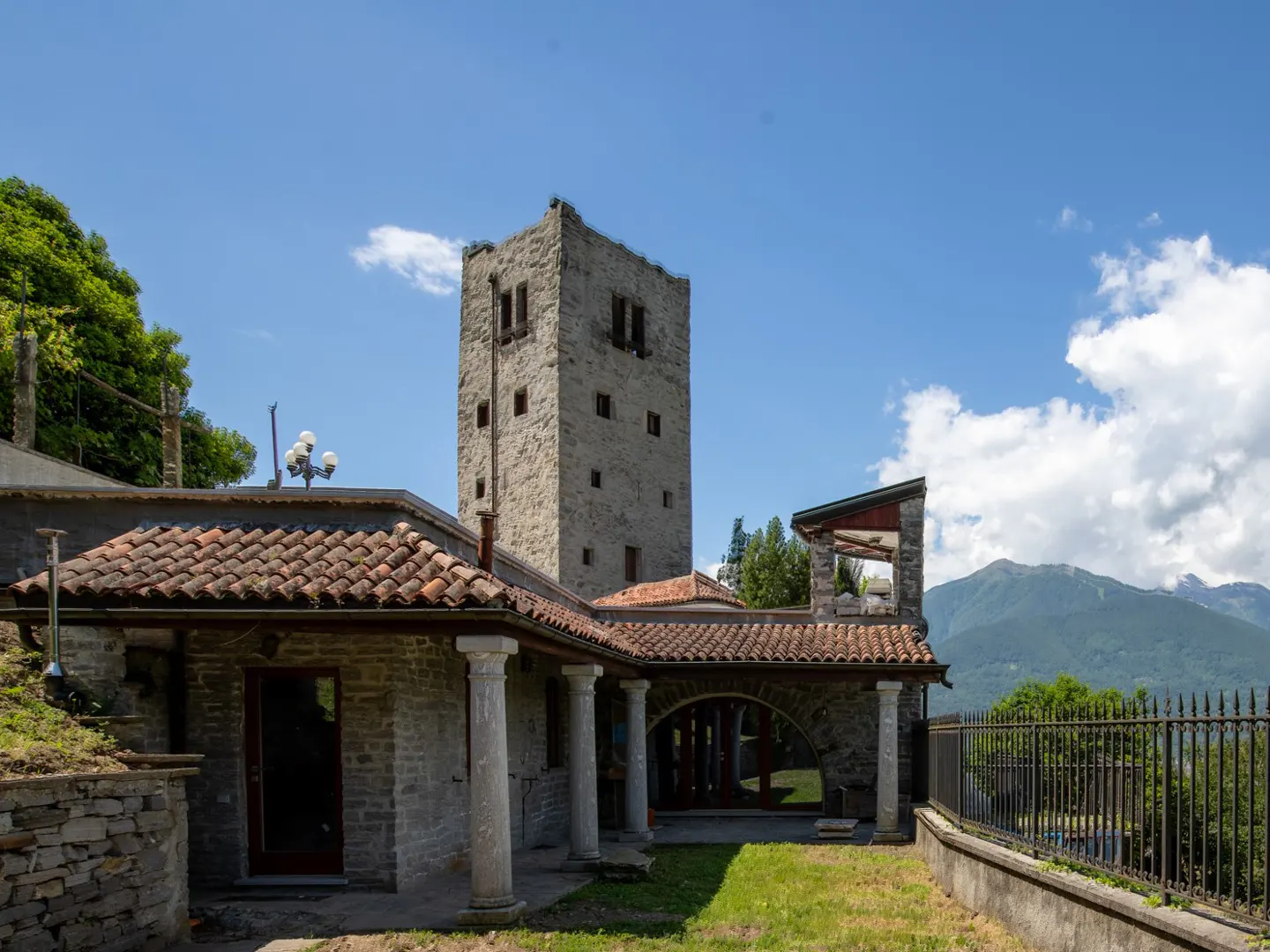 Stone building with a tower, red tile roof, and columns under a blue sky with mountains in the background.