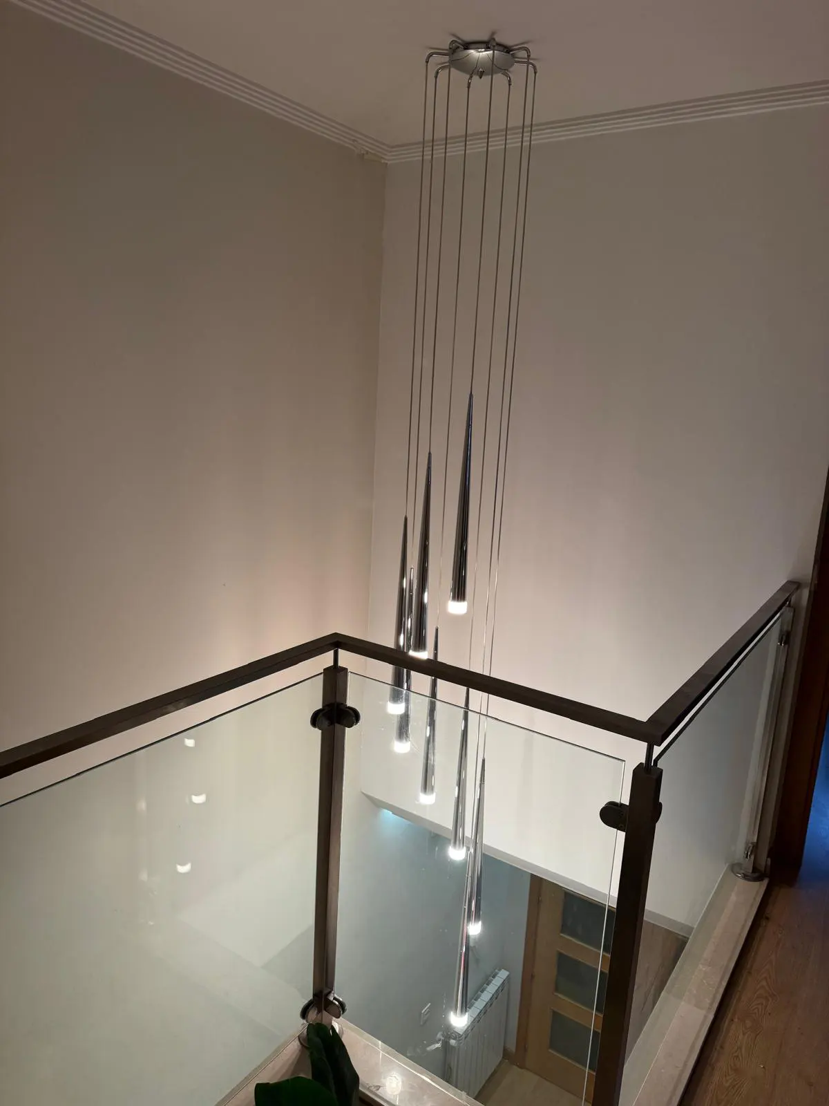 View of a modern chrome chandelier with cone-shaped lights hanging above a glass-paneled staircase railing.
