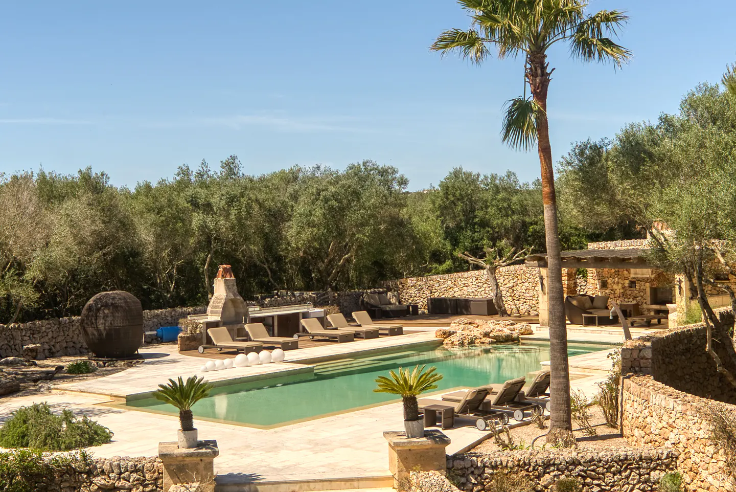 Outdoor pool area with lounge chairs, stone walls, and a palm tree under a clear blue sky.