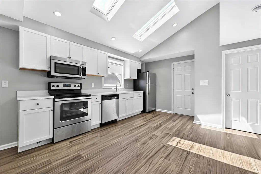 Bright kitchen with skylights, gray walls, and wood floors. White cabinets, stainless steel appliances, and two white doors are visible.