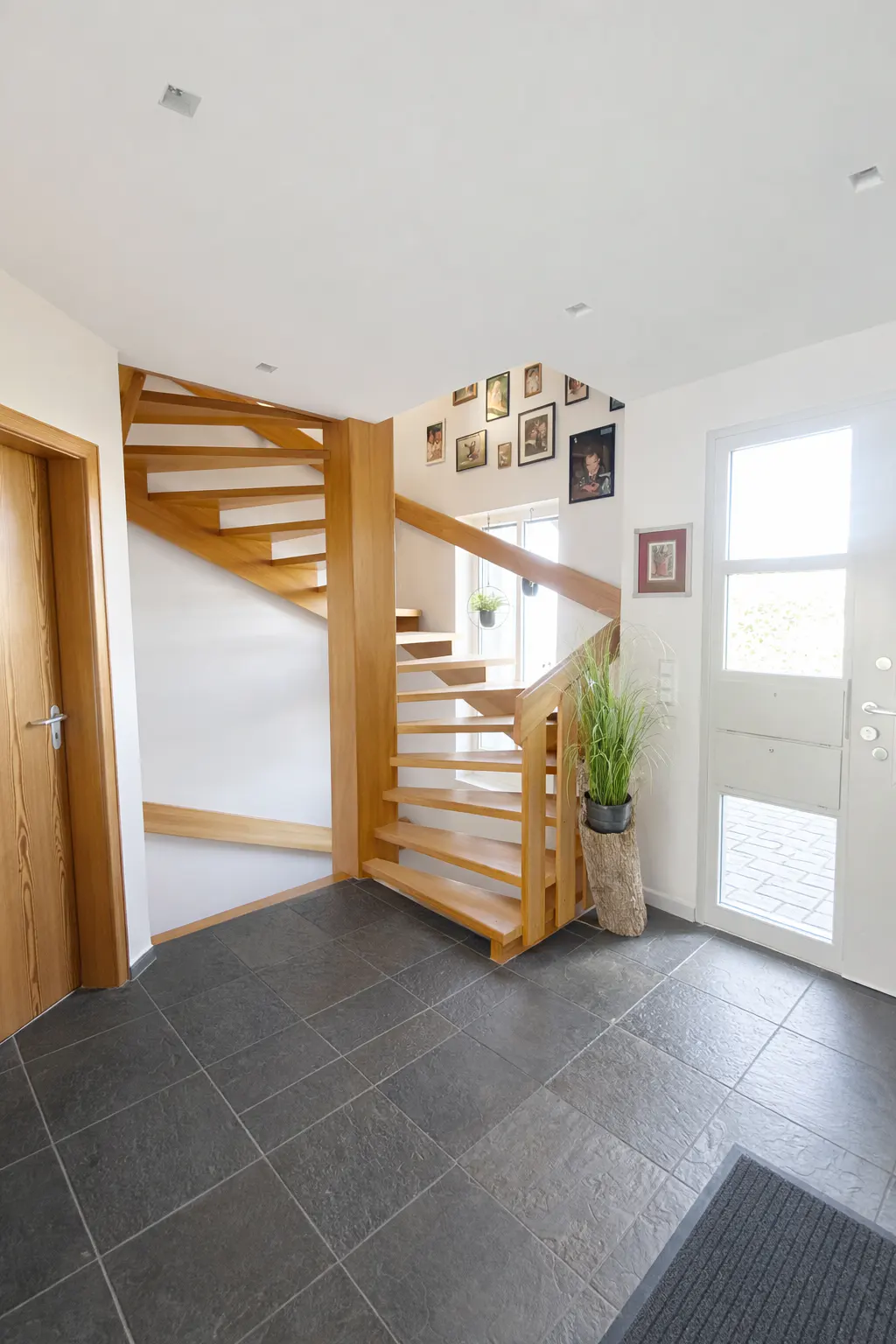 Bright foyer with a wooden staircase, gray tile floor, and white walls. A door and potted plant are visible.