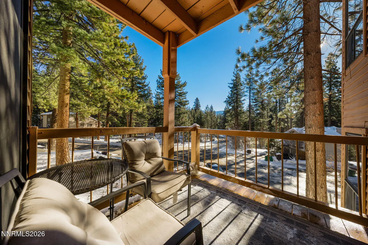 A wooden deck with chairs and a table overlooks a snowy landscape with tall pine trees under a clear blue sky.