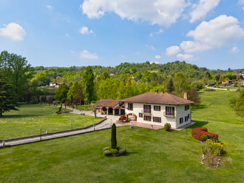 Aerial view of a cream-colored house with a brown roof, surrounded by green grass and trees under a blue sky with white clouds.