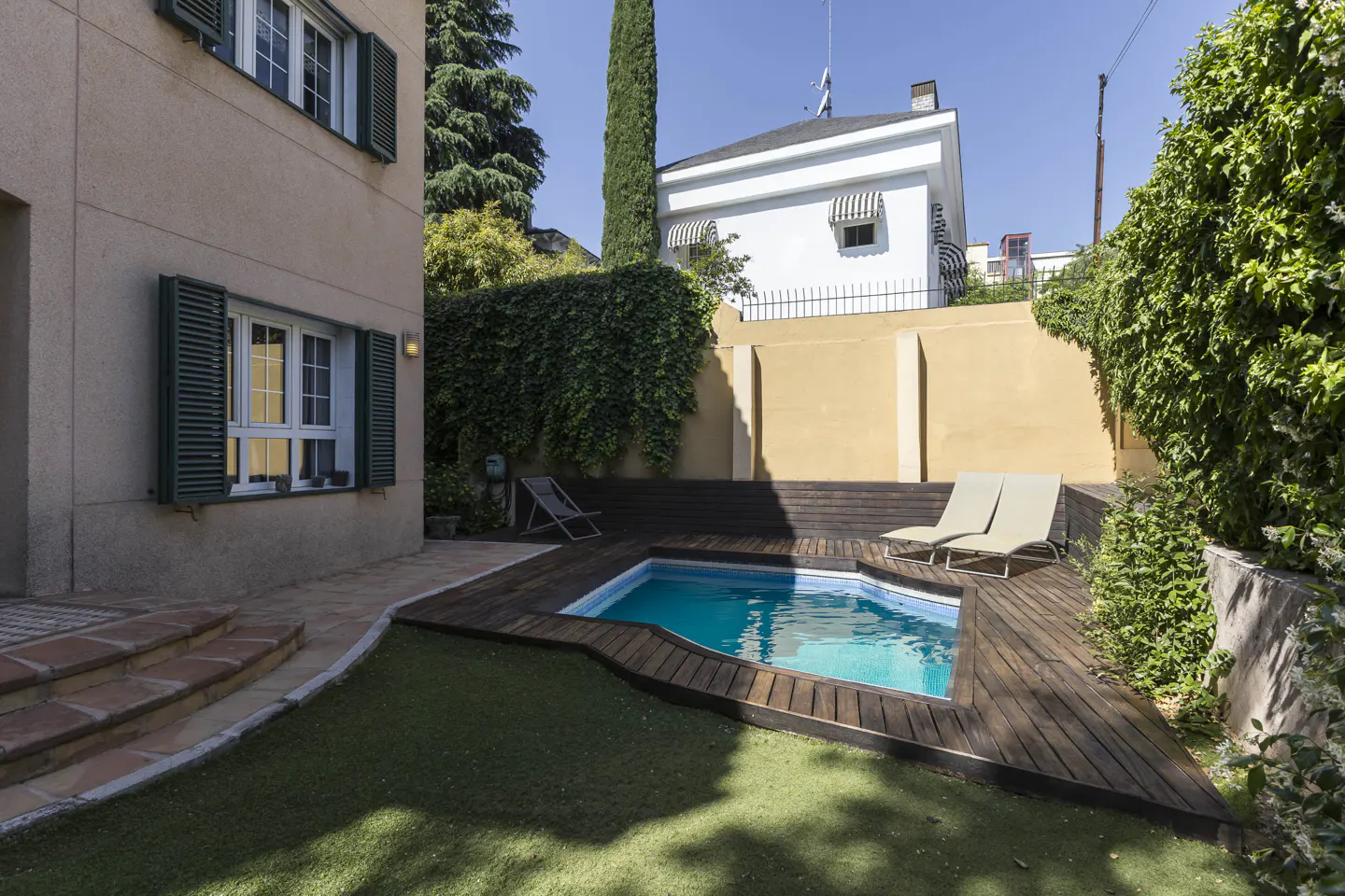 Backyard view of a small pool with a wooden deck, two lounge chairs, and green grass. A house with green shutters is on the left.
