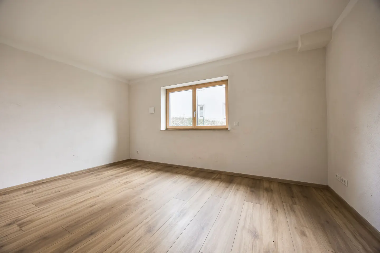 Bright, empty room with light wood floors, white walls, and a wood-framed window showing greenery outside.