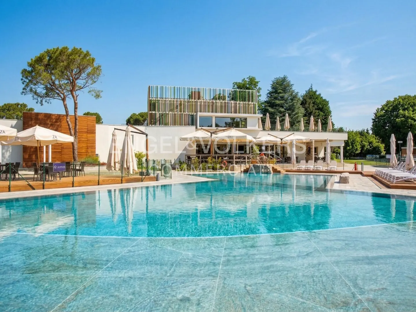 A turquoise pool reflects a modern building with umbrellas and trees under a clear blue sky.