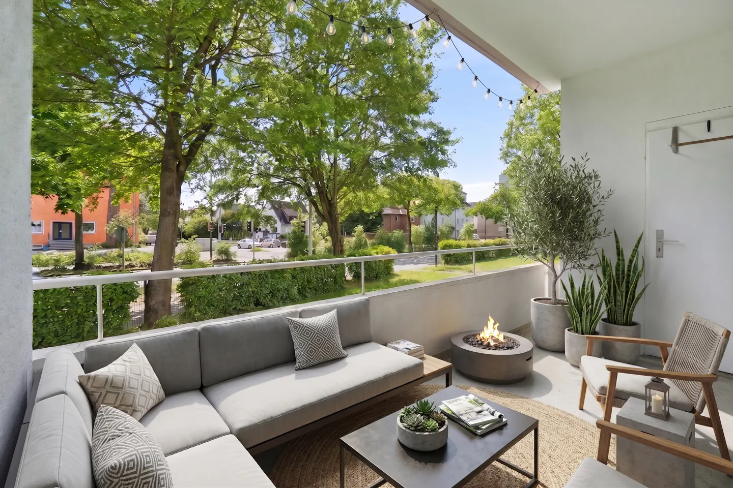 A modern balcony with gray sectional sofa, fire pit, and potted plants. String lights hang above, with a view of green trees and a street.