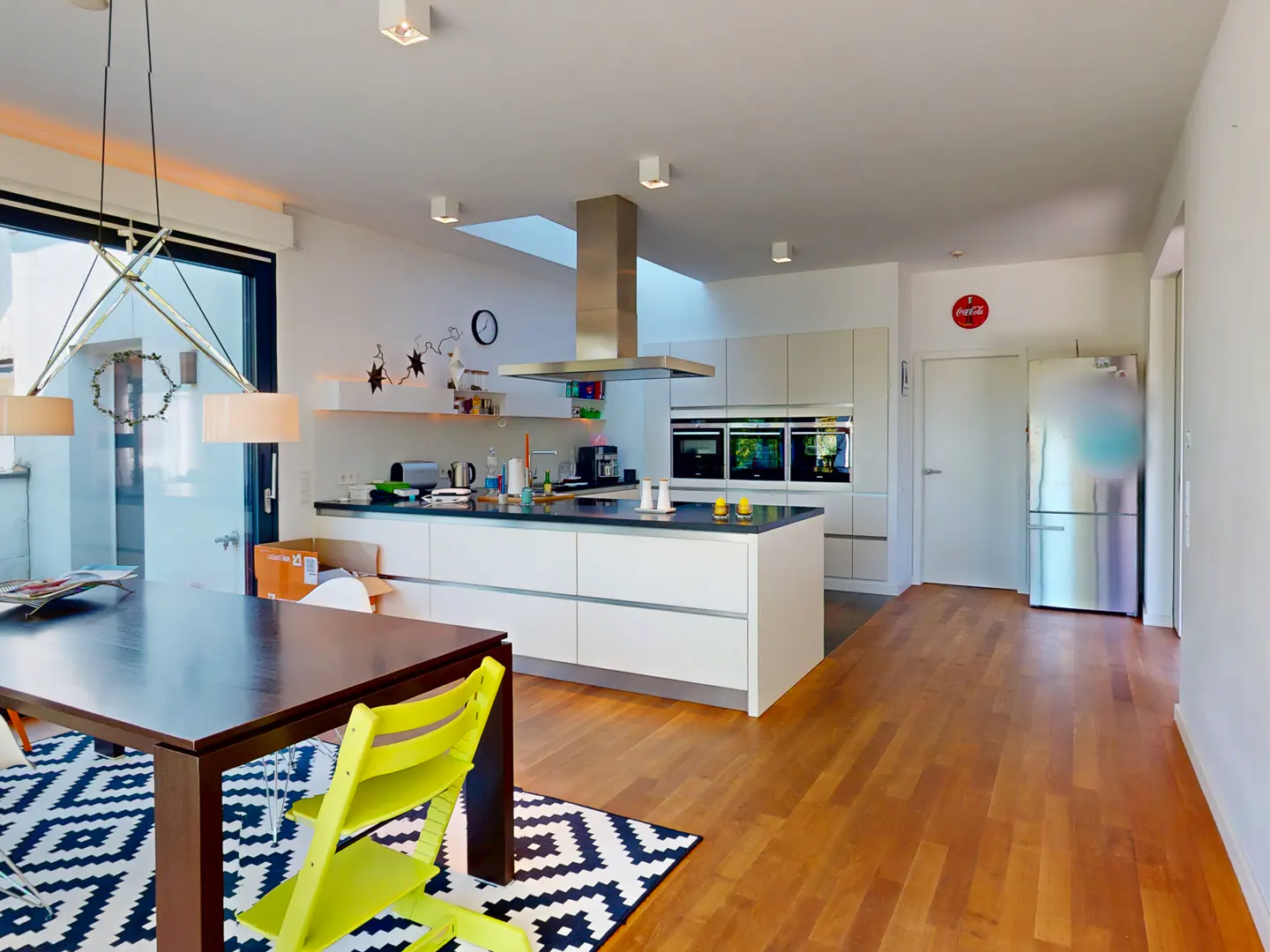 A modern kitchen with white cabinets, stainless steel appliances, and a dark wood table with a yellow chair on a black and white rug.