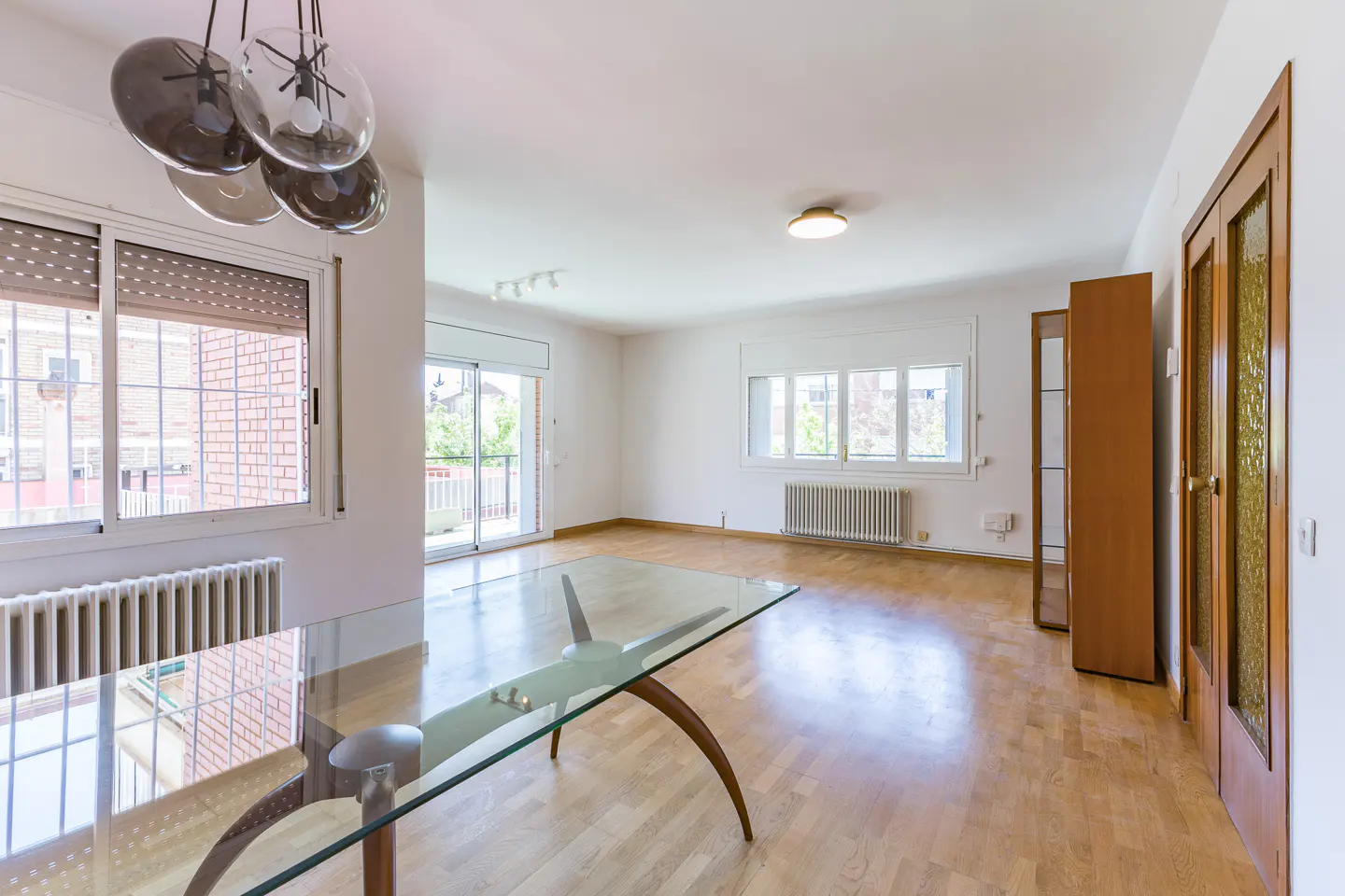 Bright, empty room with wood floors, white walls, and a glass table. Windows and a sliding glass door let in natural light. A modern chandelier hangs above.