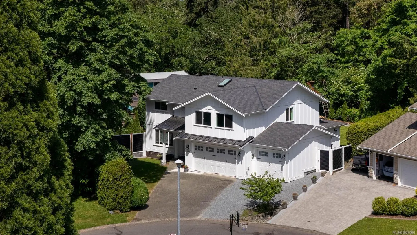 Aerial view of a two-story white house with a gray roof, surrounded by green trees. A driveway leads to a two-car garage.