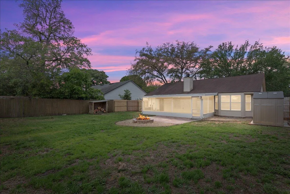 Backyard at dusk with a fire pit, green grass, a wood shed, and a screened-in porch. The sky is pink and purple.