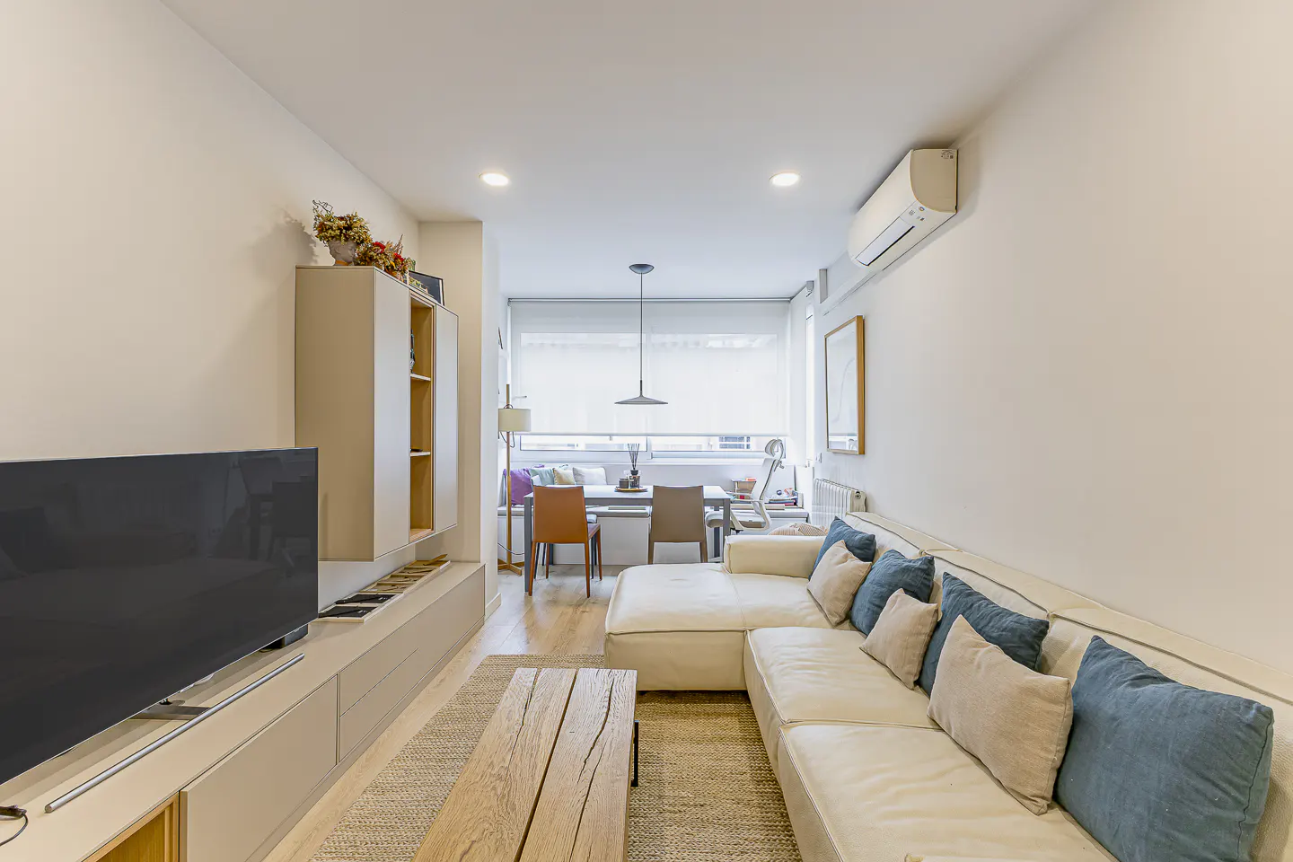 Modern living room with a white sectional sofa, blue pillows, wooden coffee table, and a dining area in the background.