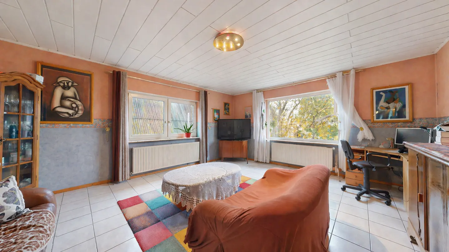 A cozy living room with peach walls, white ceiling, and tiled floor. A covered sofa, colorful rug, desk, and windows are visible.