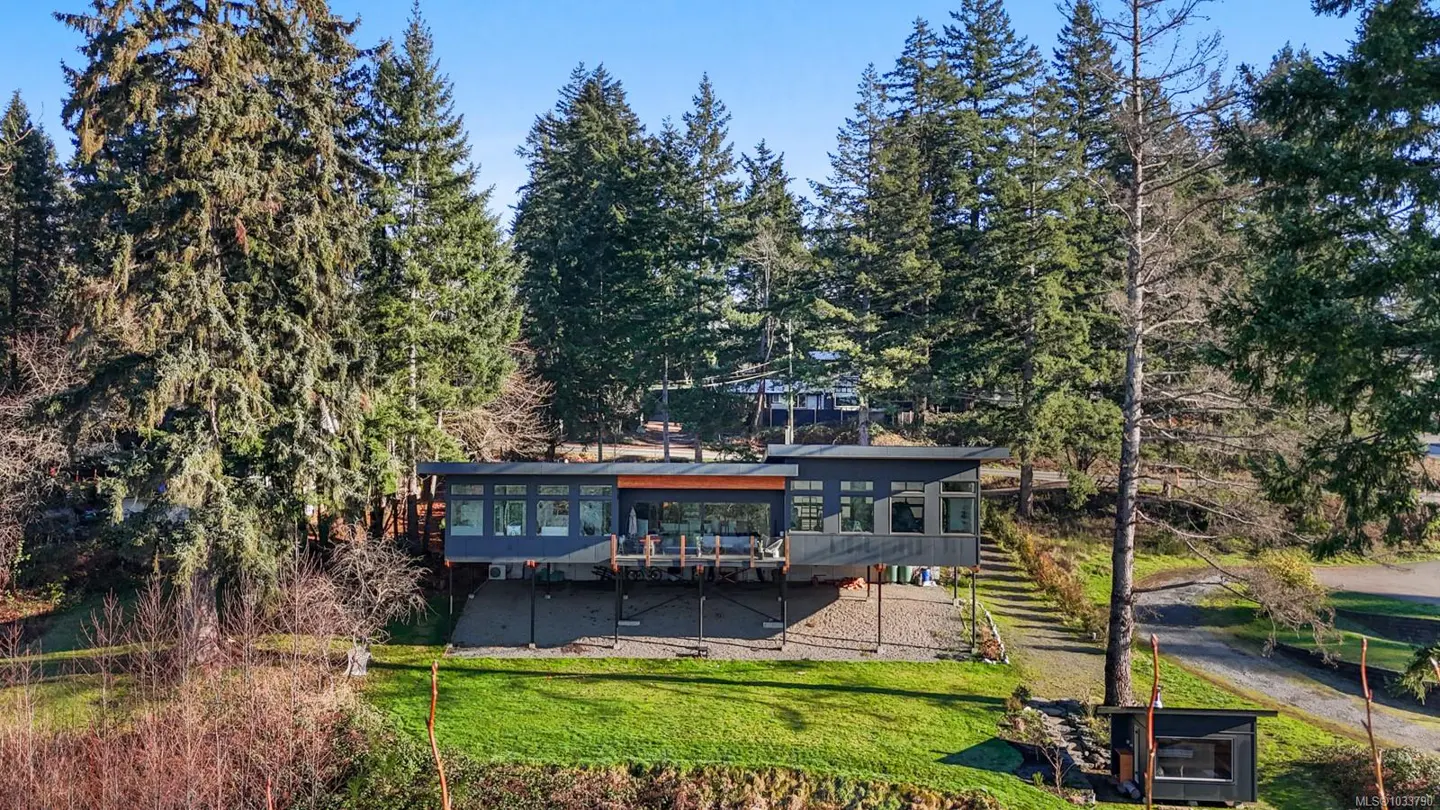 Modern dark gray house on stilts, surrounded by tall green trees and a grassy lawn under a blue sky.