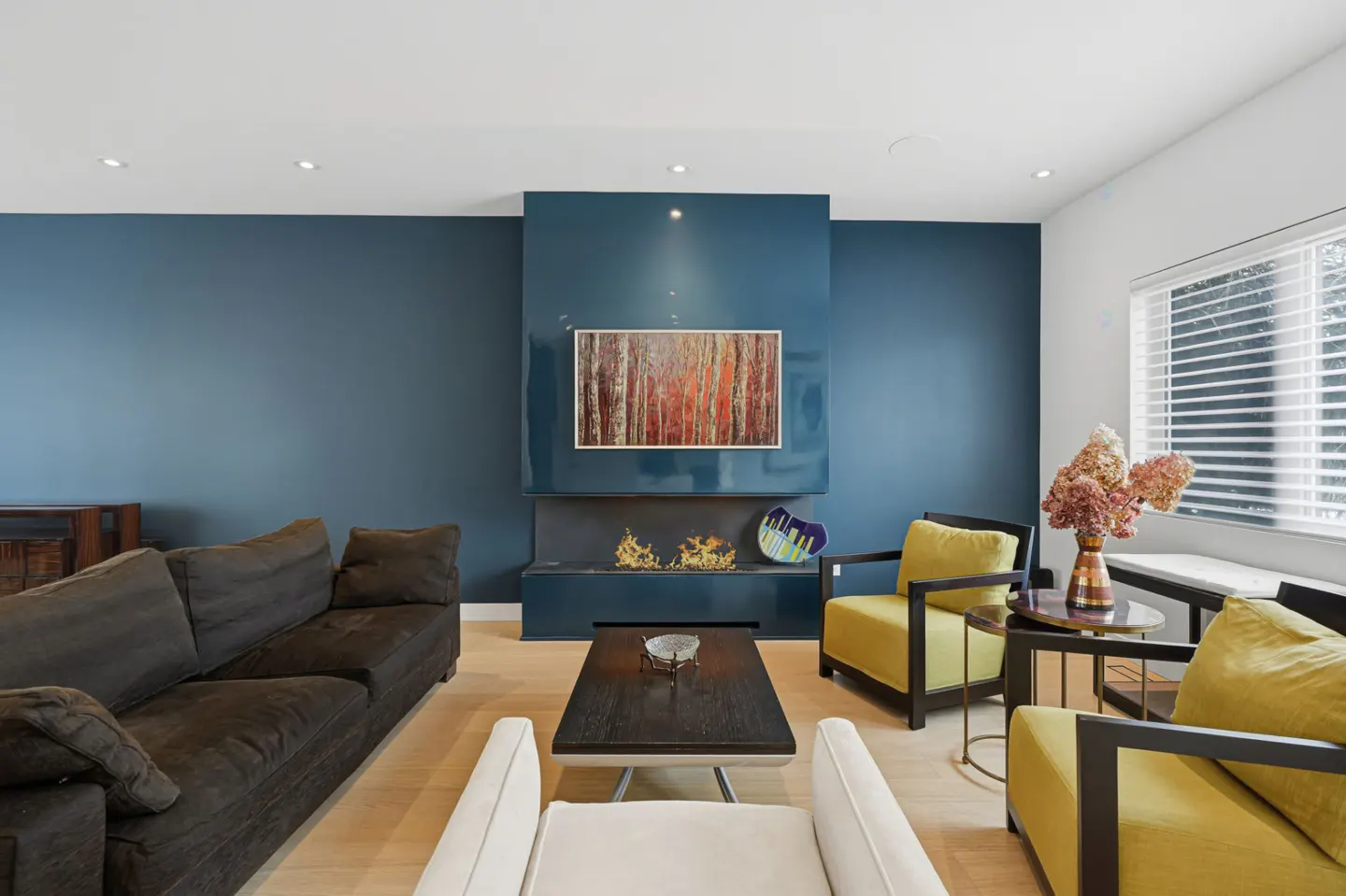 Living room with blue walls, a dark sofa, yellow armchairs, and a modern fireplace with a painting above it.