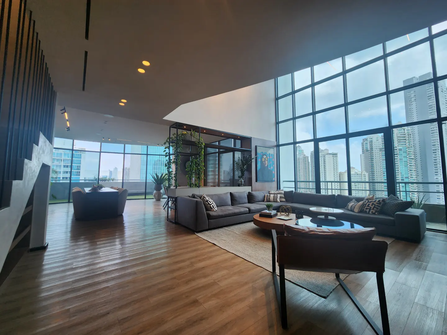 Modern living room with floor-to-ceiling windows showcasing a city view. Gray sofas, wood floors, and a round coffee table create a stylish space.