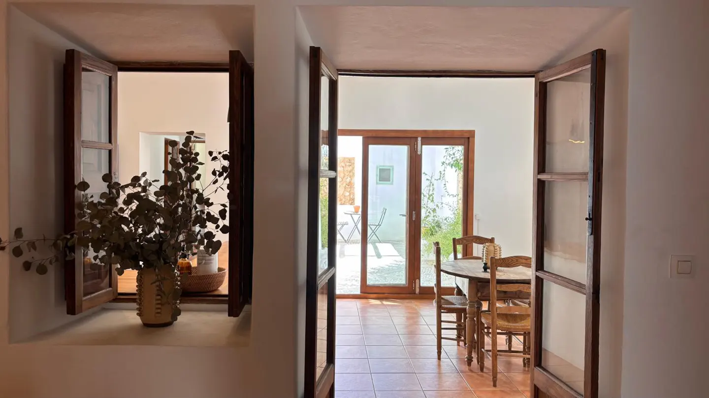 Interior view through open wood-framed doors and window. A vase of eucalyptus sits on the window sill. A dining table and chairs are visible in the next room.
