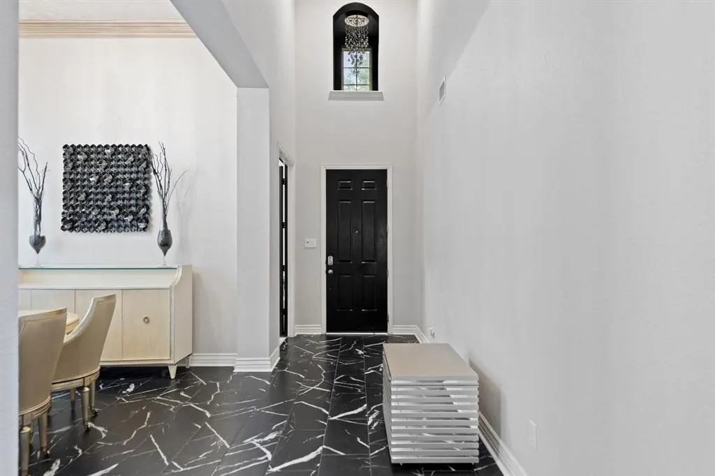 A bright foyer with black marble floors leads to a black front door. A dining area is to the left, and a silver bench is to the right.
