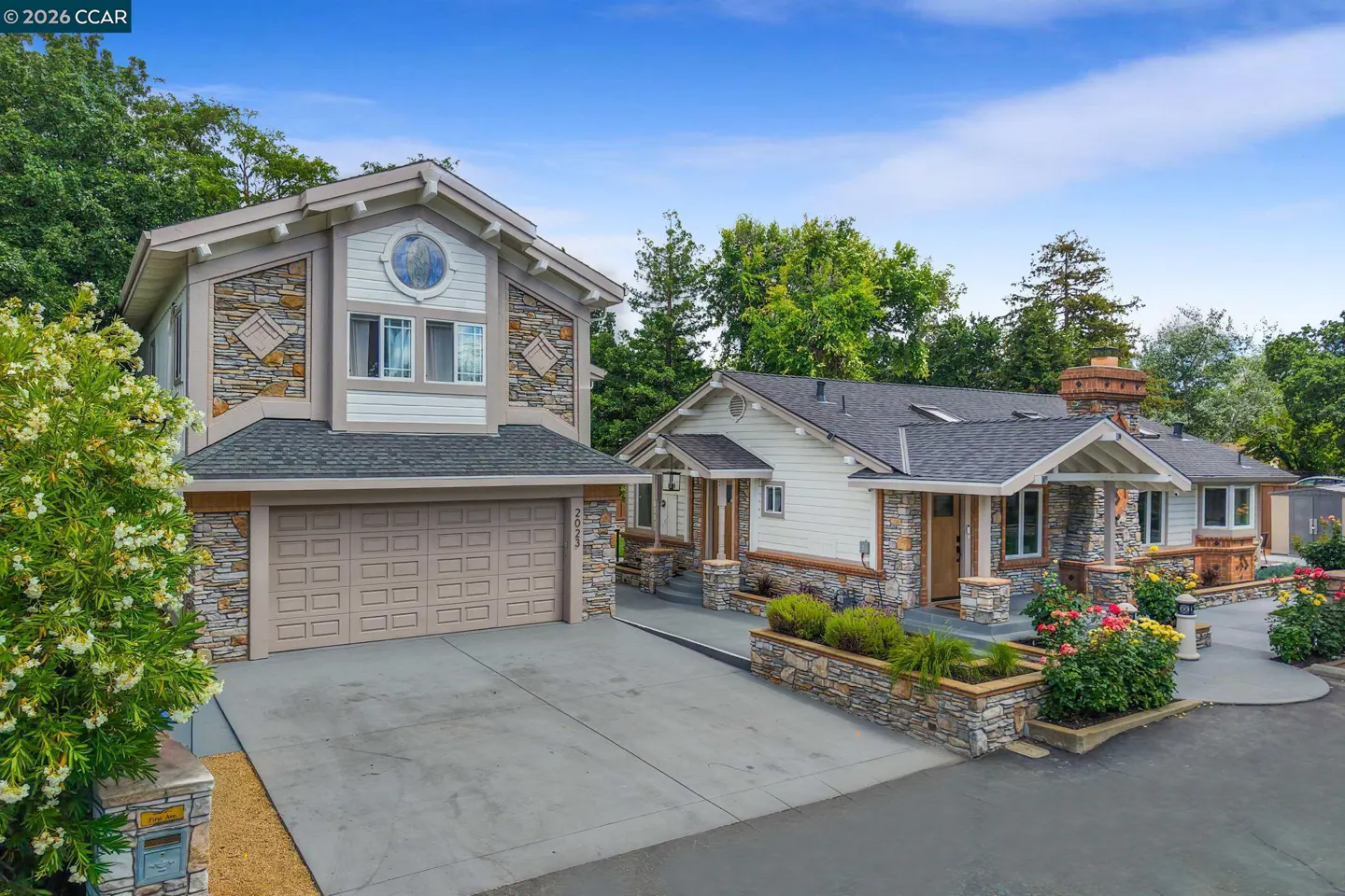 Exterior view of a two-story house with a beige garage door, stone accents, and a gray roof, surrounded by green trees.