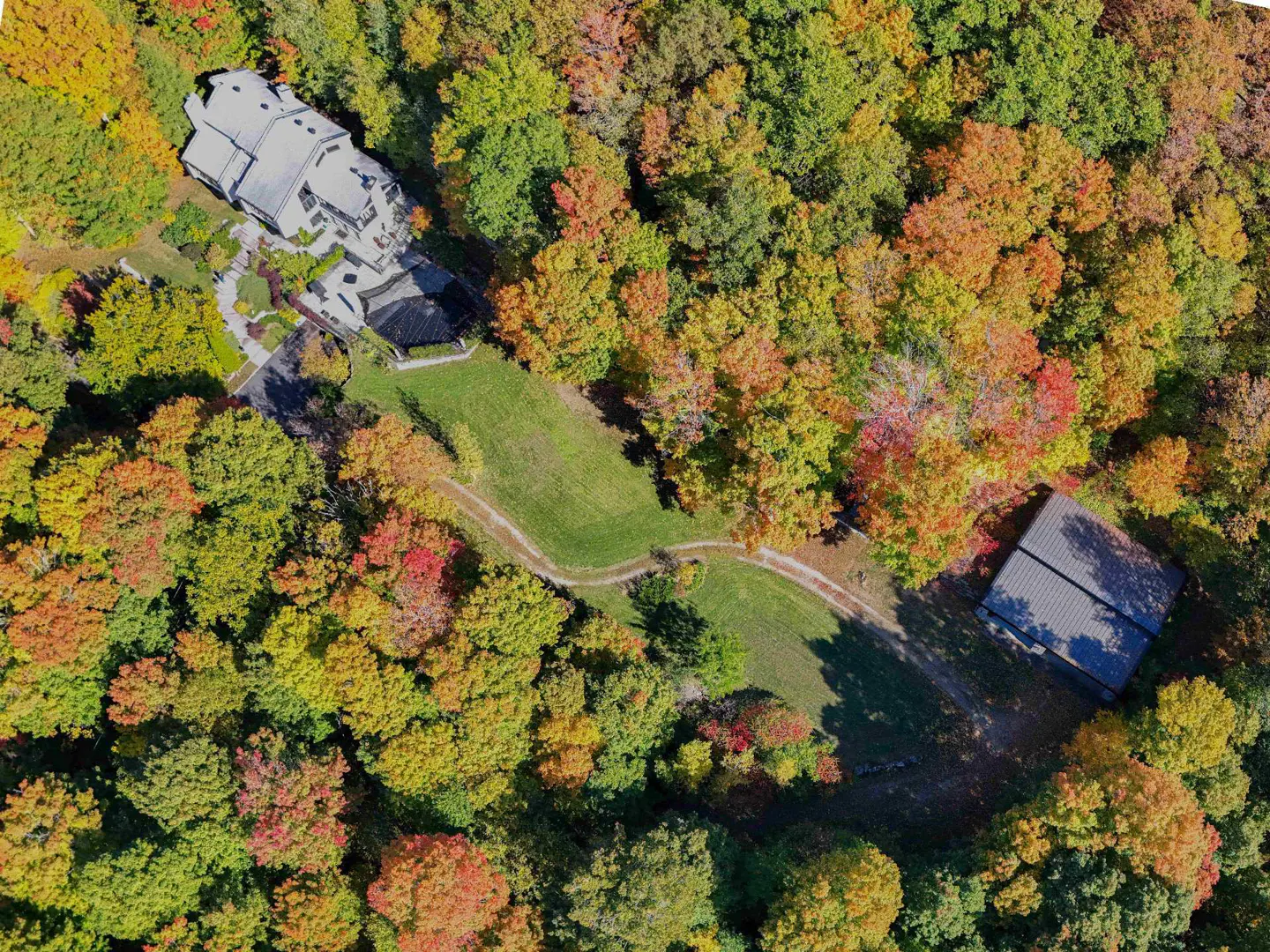 Aerial view of a gray house and a shed surrounded by colorful autumn trees.