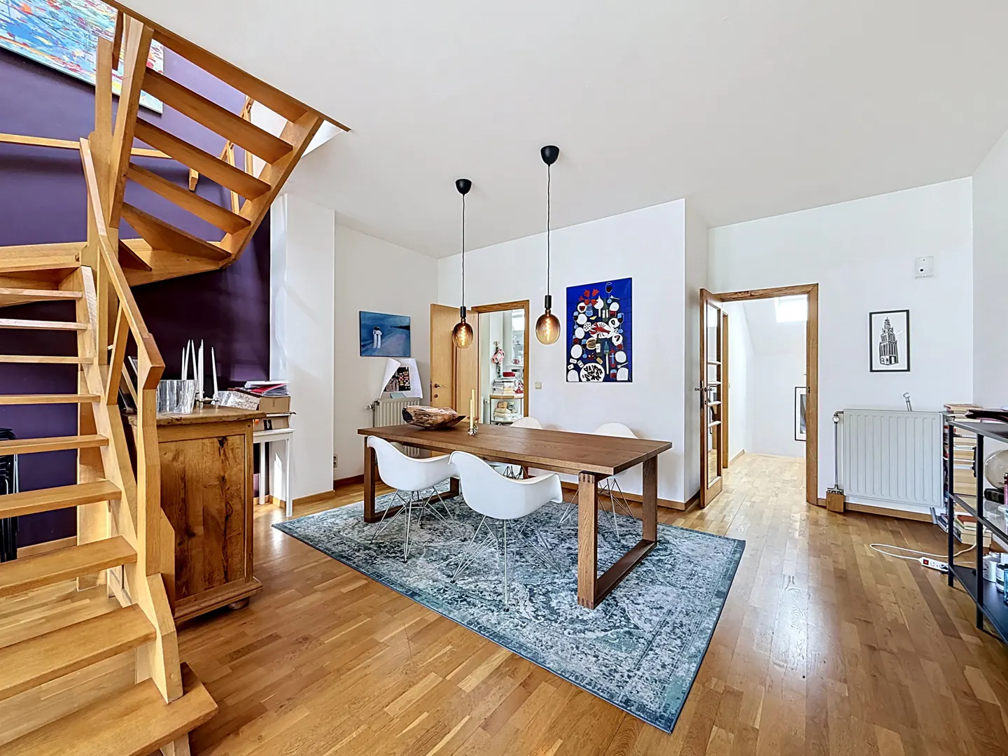 Dining room with wooden table, white chairs, blue patterned rug, and a wooden staircase against a deep purple wall.