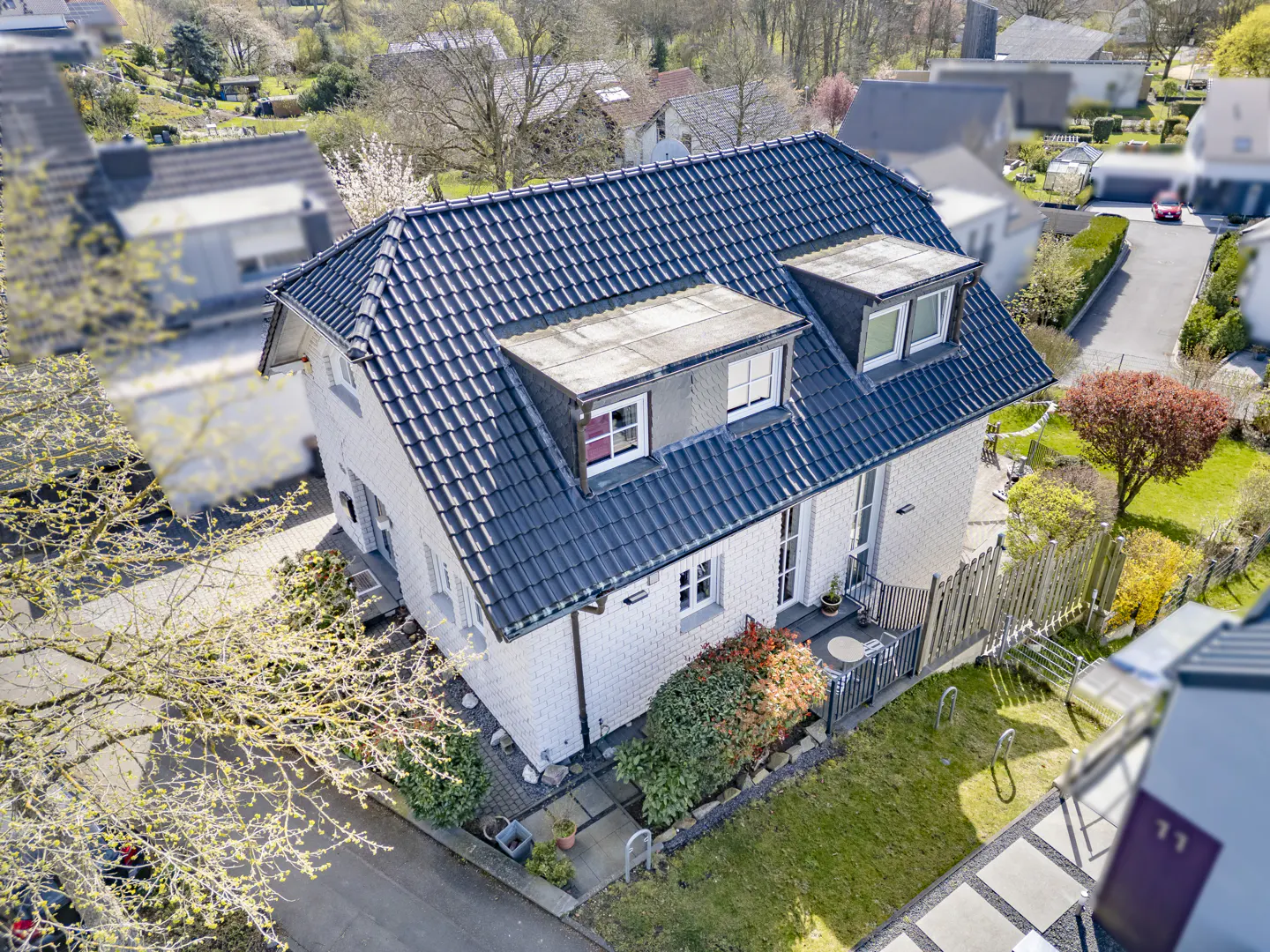 Aerial view of a two-story white brick house with a dark blue tiled roof and dormer windows, surrounded by green lawns and trees.