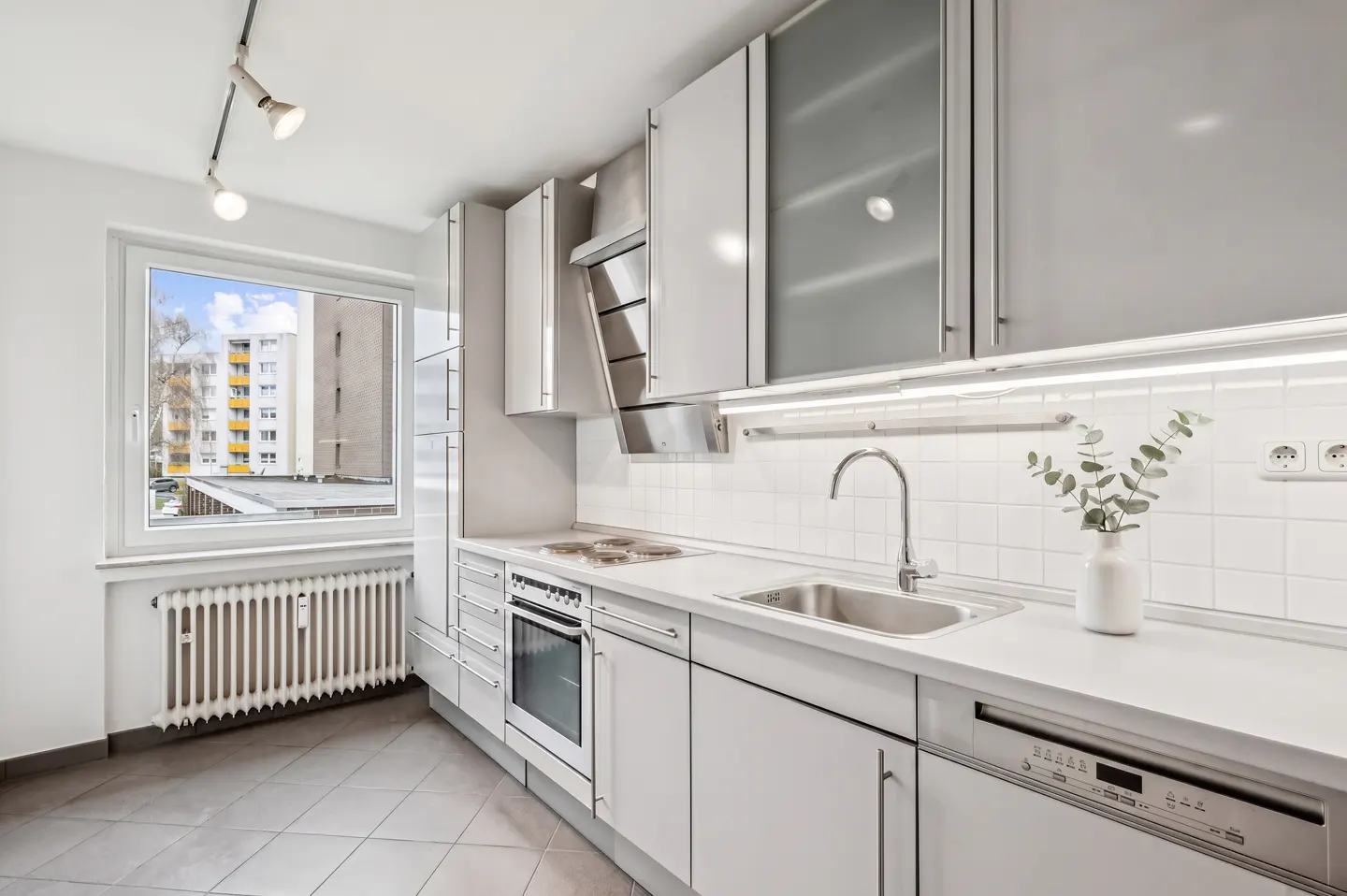 Bright kitchen with white cabinets, stainless steel appliances, and a window overlooking a building. Gray tile floor.