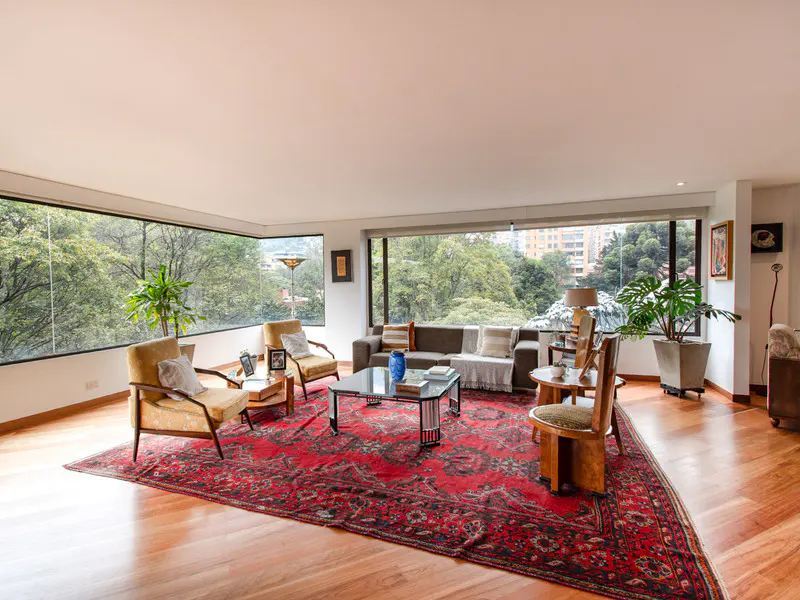 Bright living room with wood floors, a red patterned rug, and large windows overlooking lush greenery. A brown sofa and several chairs furnish the space.