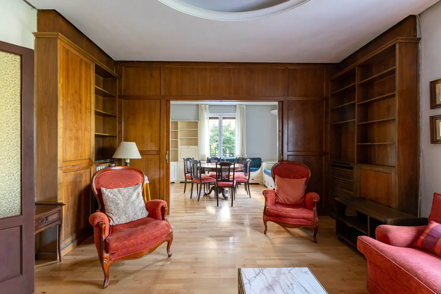 Living room with wood paneling and built-in bookshelves. Red armchairs and sofa. Dining room visible through doorway.