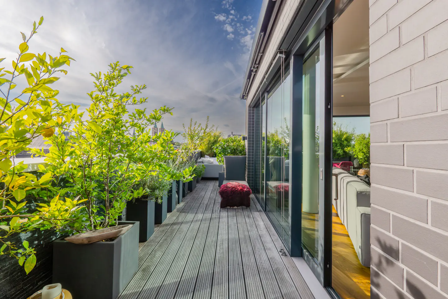A modern balcony with potted plants, a wooden floor, and a view of the city skyline under a blue sky. Sliding glass doors lead to an interior living space.