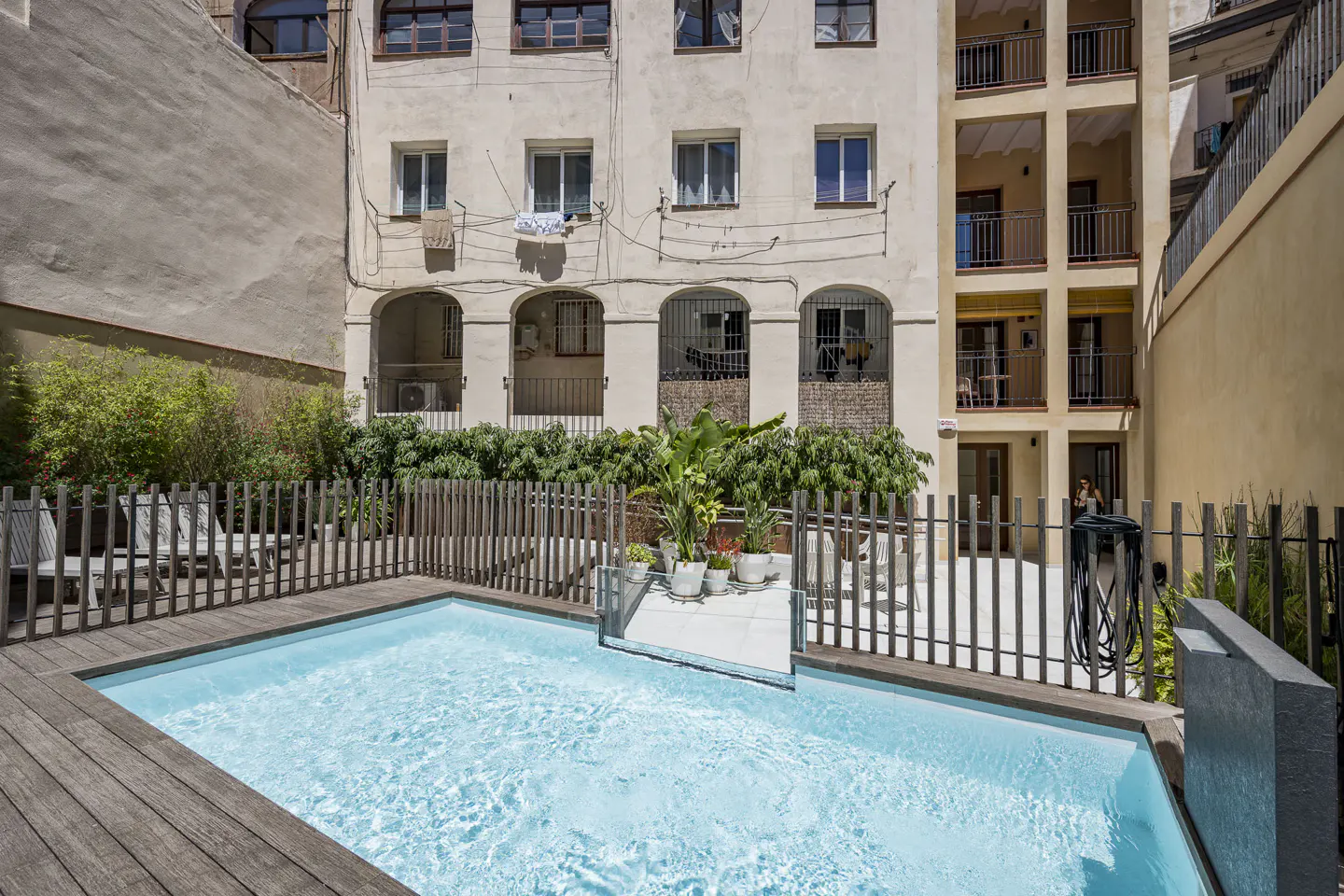 A rectangular pool with blue water is surrounded by a wooden deck and fence in a courtyard. Tall buildings are in the background.