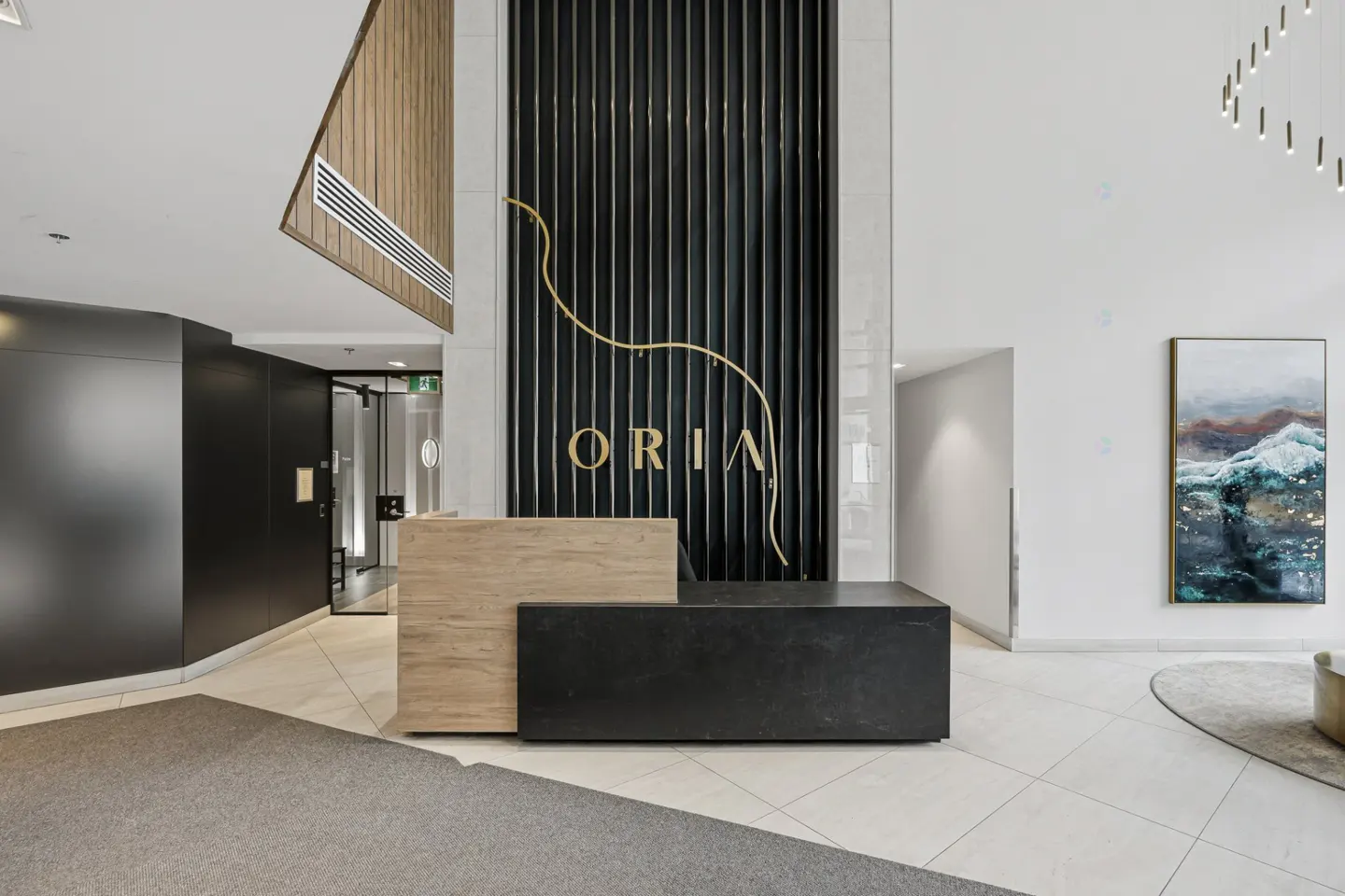 Modern lobby with a black and light wood reception desk. "ORIA" is displayed on a black slat wall with a gold design. A painting hangs on the white wall.
