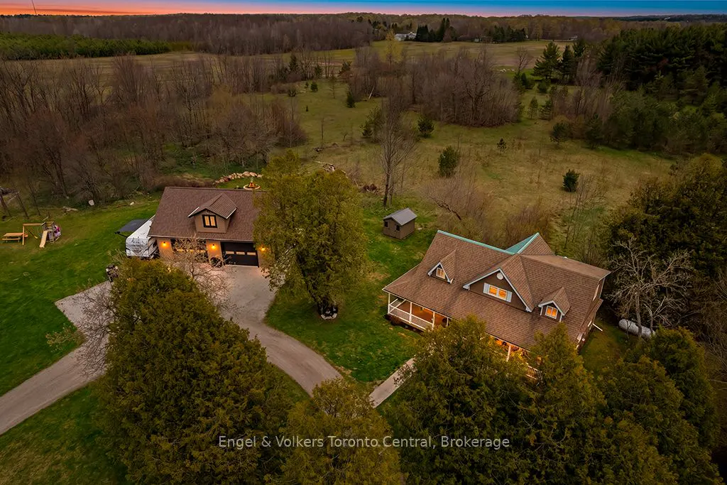 Aerial view of two brown houses with green lawns and trees, set against a backdrop of fields and forest under a twilight sky.