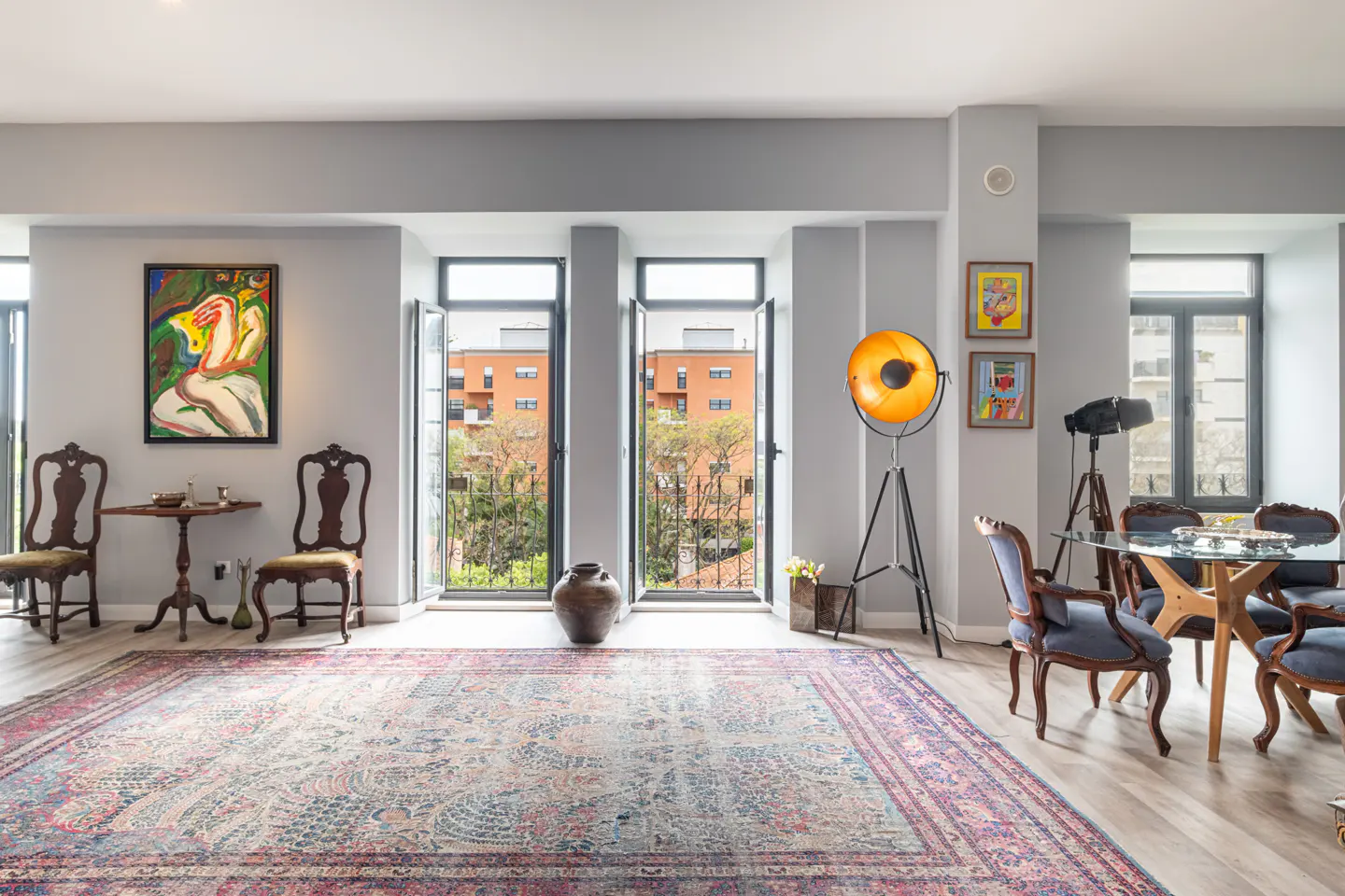 A bright living room with a patterned rug, gray walls, and open windows. There is a dining table with blue chairs and a large yellow lamp.