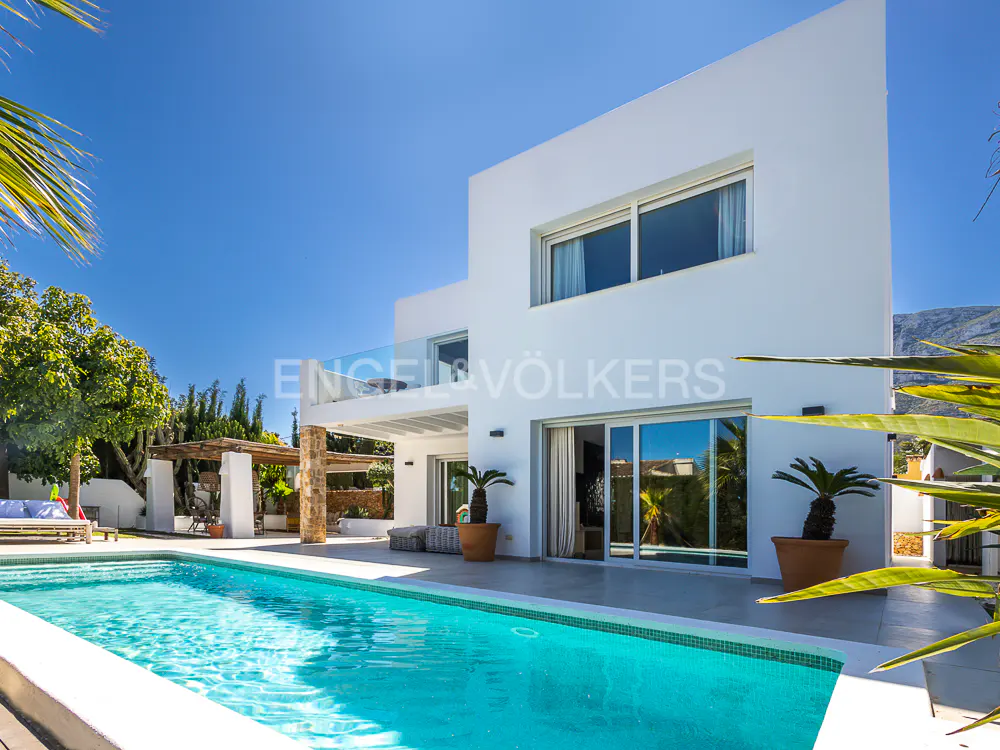 Exterior view of a modern white house with a blue swimming pool in the foreground on a sunny day.