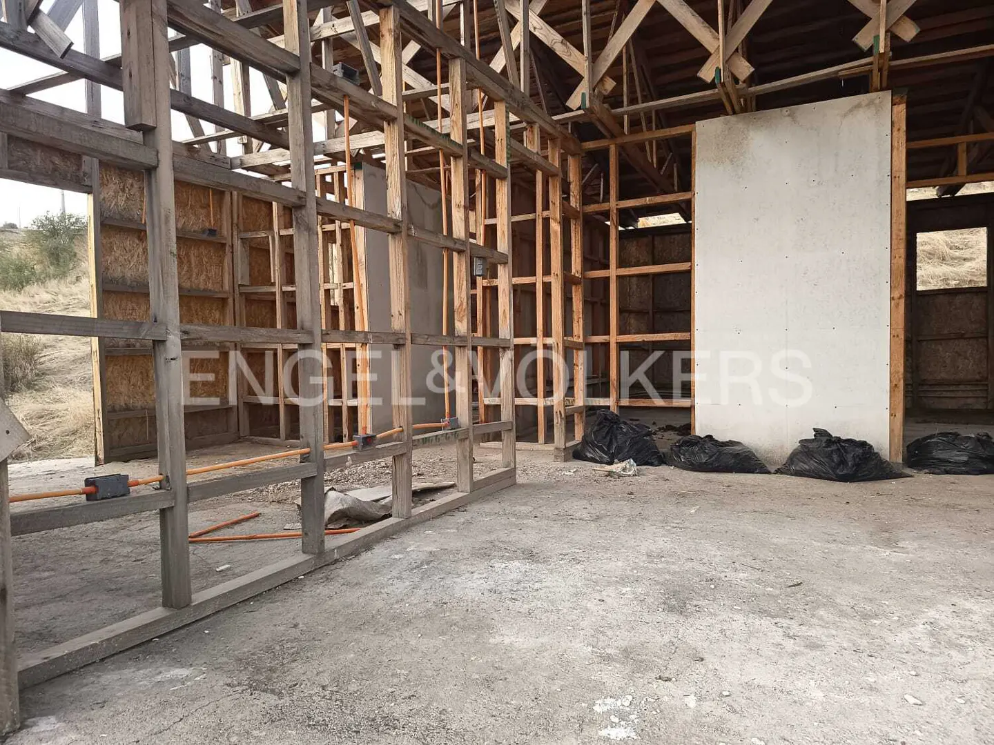 Interior view of a building under construction with exposed wooden beams and black trash bags on the concrete floor.