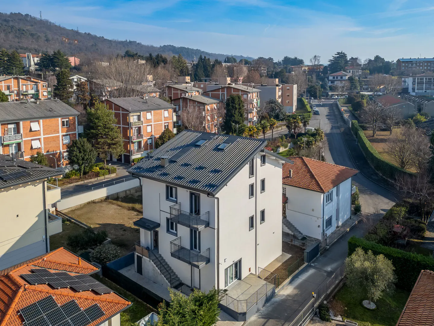 Aerial view of a modern, white three-story house with a gray roof and black balconies in a suburban neighborhood on a sunny day.