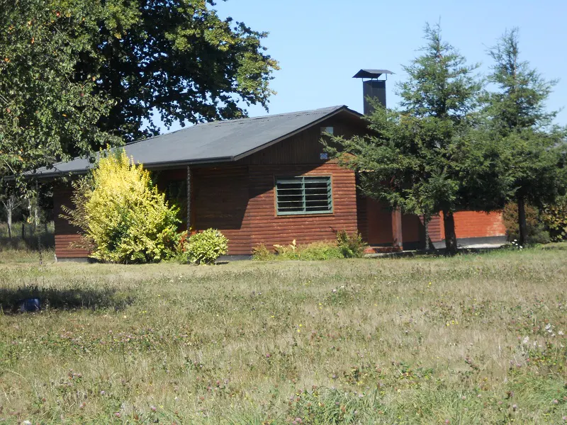 A brown, one-story house with a gray roof, chimney, and green window sits on a grassy lawn.