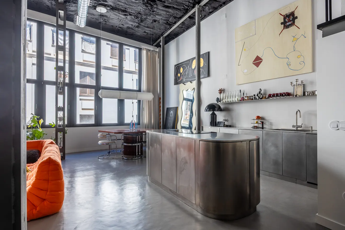 Modern loft apartment with stainless steel kitchen island, art, and large windows. Orange sofa in the foreground.