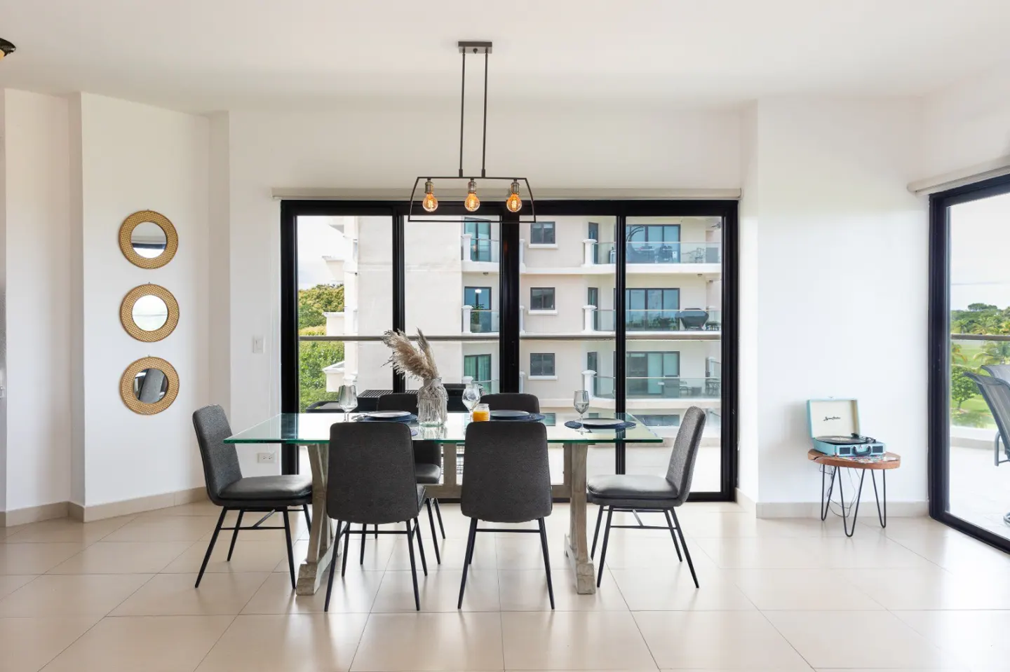 Bright dining room with glass table, gray chairs, and black-framed windows overlooking a building. Three round mirrors hang on the wall.